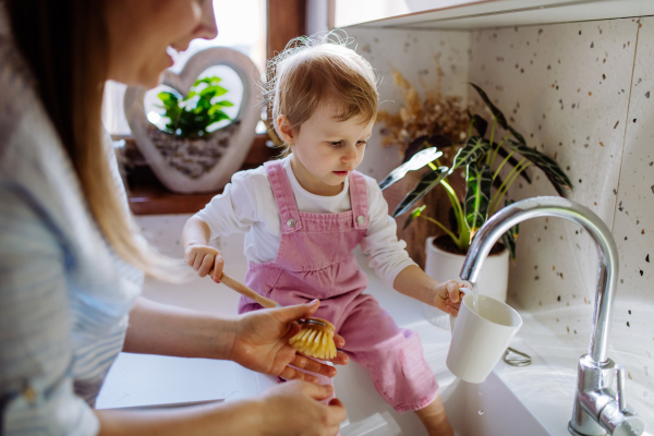 Little girl sitting on kitchen counter and helping mother to wash cup in sink in kitchen.