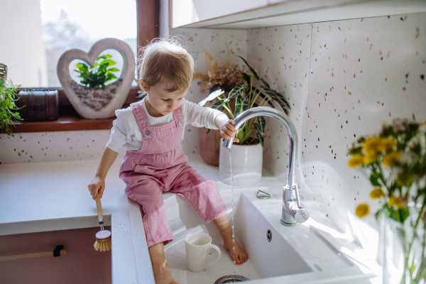 A little girl sitting on kitchen counter and washing cup in sink in kitchen.