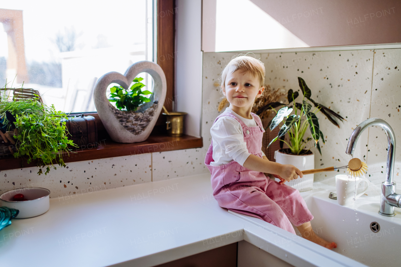 A little girl sitting on kitchen counter and washing cup in sink in kitchen.