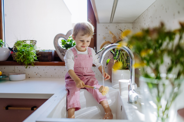 A little girl sitting on kitchen counter and washing cup in sink in kitchen.