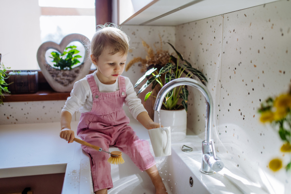 A little girl sitting on kitchen counter and washing cup in sink in kitchen.