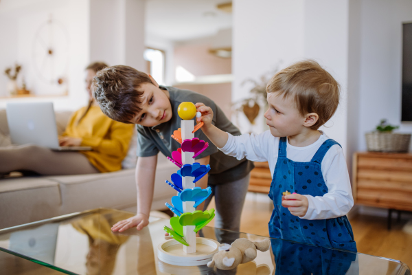 Little siblings playing with a montessori wooden marble run in living room, their mother is sitting on sofa with laptop.