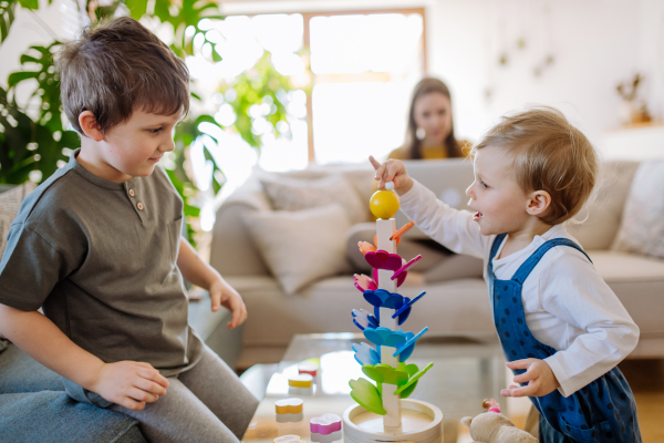 Little siblings playing with a montessori wooden marble run in living room, their mother is sitting on sofa with laptop.