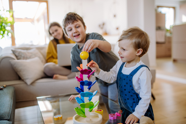 Little siblings playing with wooden toys in living room, their mother is sitting on a sofa with laptop.