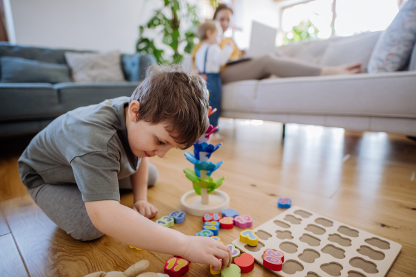 Little boy playing with montessori wooden toys in living room,mother and sister in the backgroud.