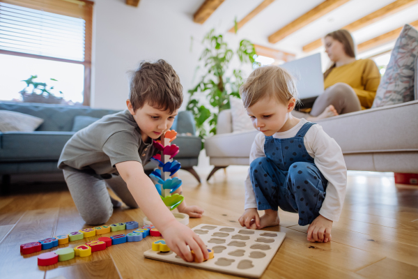 Little siblings playing with montessori wooden toys in living room, their mother is sitting on a sofa with laptop.