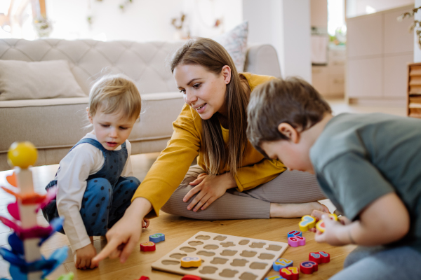 Young mother and kids playing with montessori wooden toys in living room, having nice family time together.