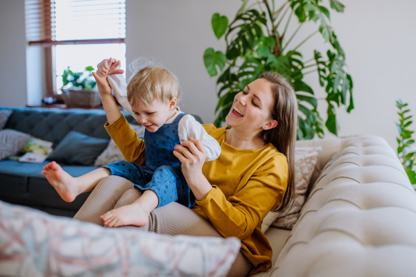 A young cheerful mother playing with her little child and having fun when lifting her up on sofa.