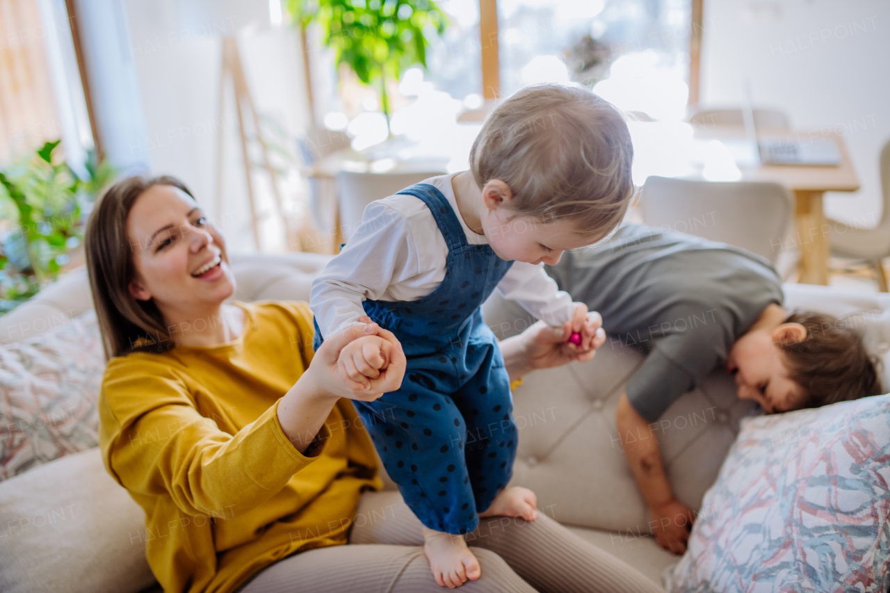 A young cheerful mother playing with her little children and having fun when lifting them up on sofa.