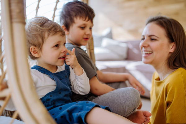 A young cheerful mother playing with her little children and having fun when swinging them on hanging chair in conservatory at home.