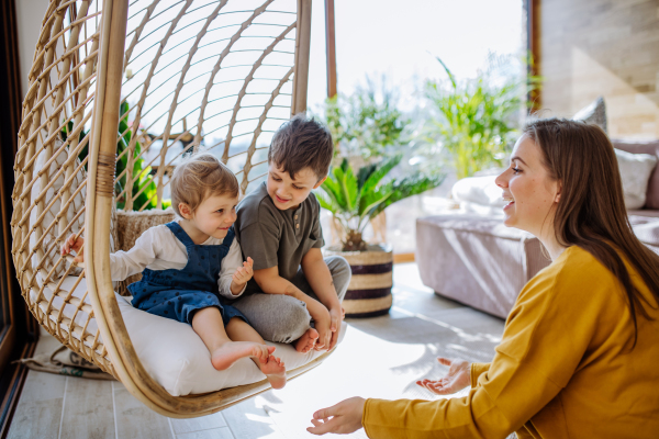 A young cheerful mother playing with her little children and having fun when swinging them on hanging chair in conservatory at home.