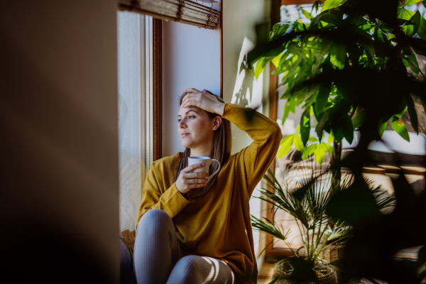 A portrait of happy young woman resting and daydreaming at home, shot through window.