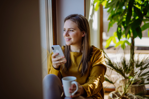 Young college student using phone, when sitting on floor at home and enjoying a cup of tea.