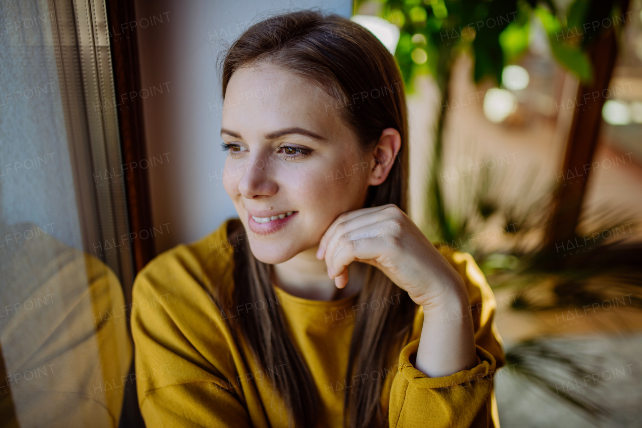 A portrait of happy young woman resting and daydreaming at home,looking through window