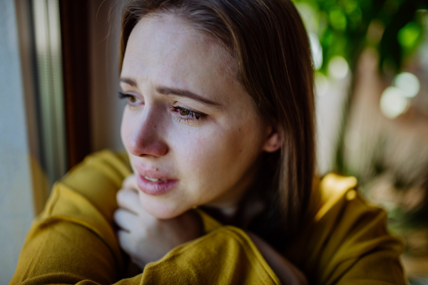 Young woman suffering from depression at home, crying and looking trough the window.