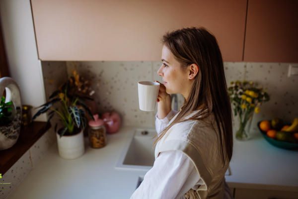 A smiling young woman with dreamy face drinking morning coffee at her kitchen.