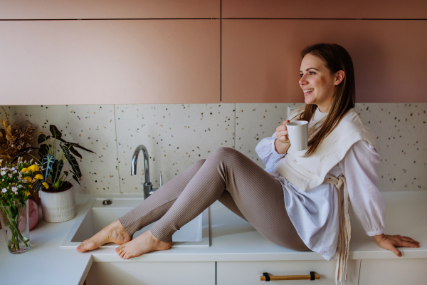 A cheerful young woman holding coffee cup while sitting on the kitchen counter