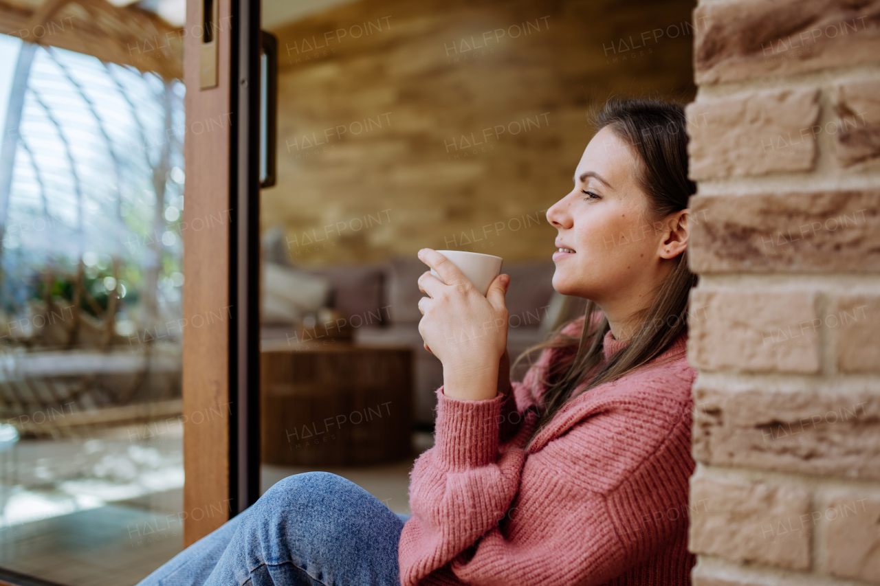 A smiling young woman with dreamy face drinking morning coffee and sitting at her home patio