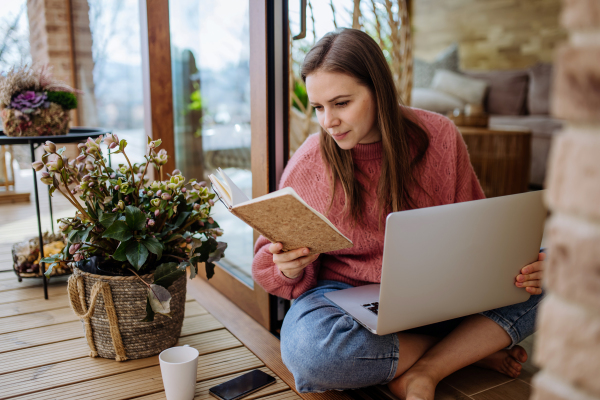 A young college student studying on laptop and using phone, when sitting on floor at home.