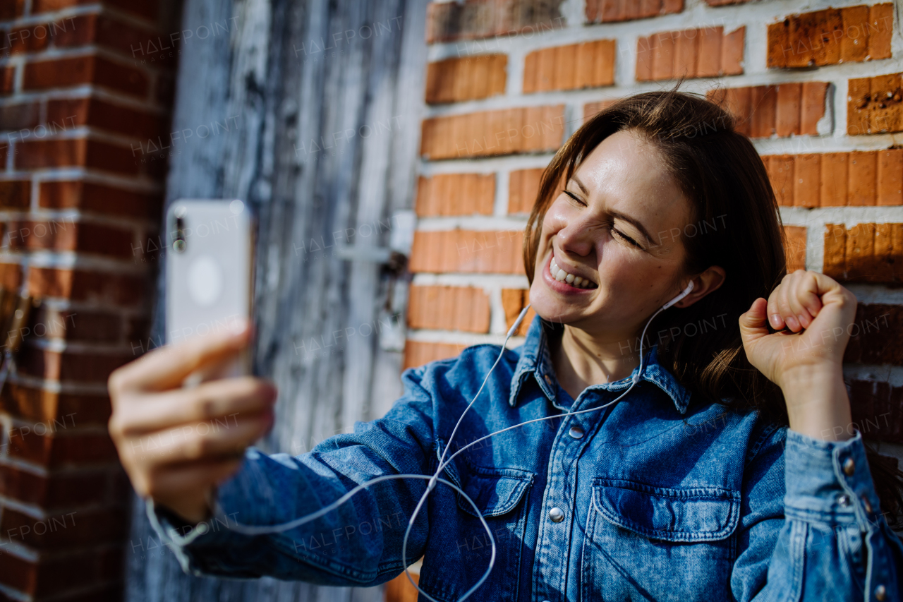 A happy young woman in denim jacket leaning the brick wall outdoors in summer and taking selfie