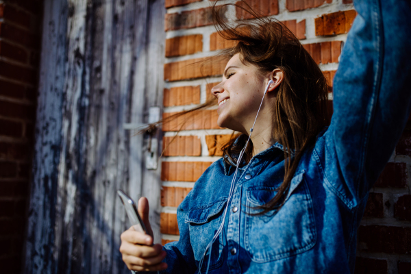 A happy young woman in denim jacket leaning the brick wall outdoors in summer and listening music from smartphone.