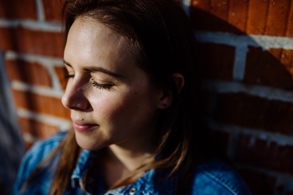 Portrait of happy young woman resting with closed eyes at home, in front of brick wall.