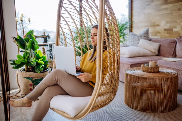 Work from home or study online, concept with a woman working at laptop outdoor on terrace, sitting in hanging chair.