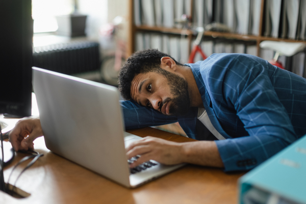 Young exhausted male employee in front of notebook in his office. Signs of burnout at work.