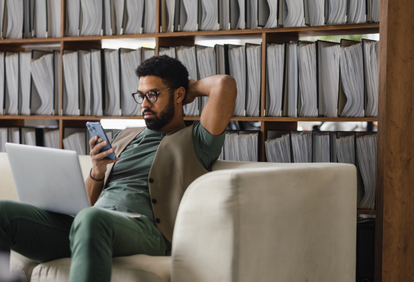 Young multiracial man sitting on a sofa and working, using notebook and smart phone in office. Manager looking at mobile phone with concerned look.