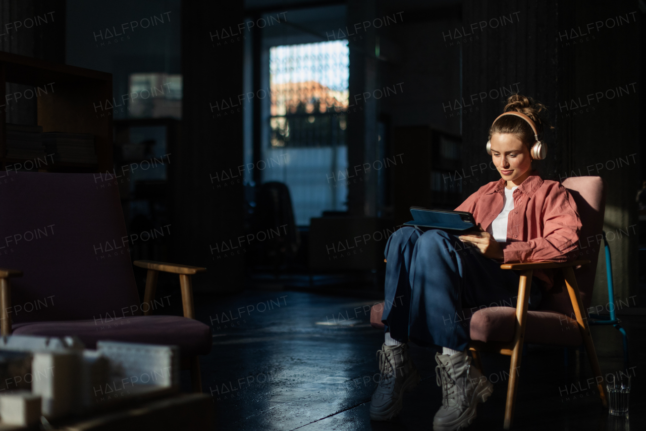 Young woman stting in armchair, listening music and working on a digital tablet.