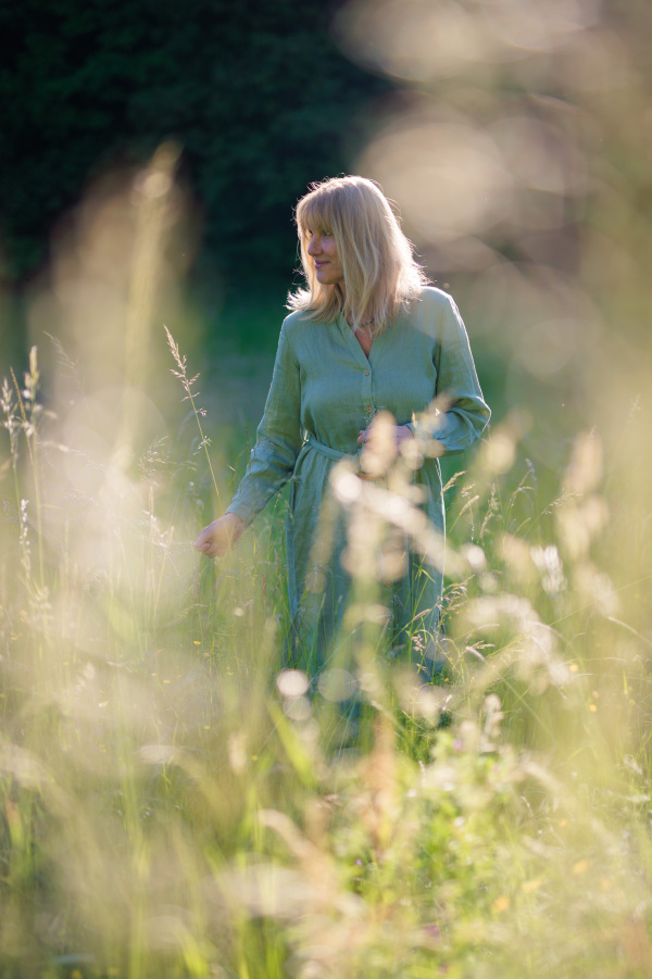 A senior woman in linen dress on walk in meadow in summer, mental health concept.