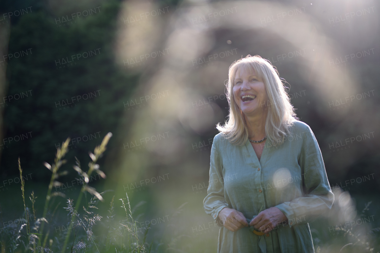 A cheerful mature woman in linen dress on walk in meadow in summer, mental health concept.