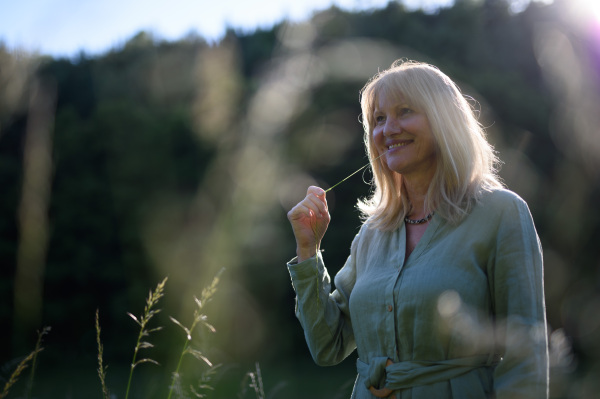 A cheerful mature woman in linen dress on walk in meadow in summer, mental health concept.