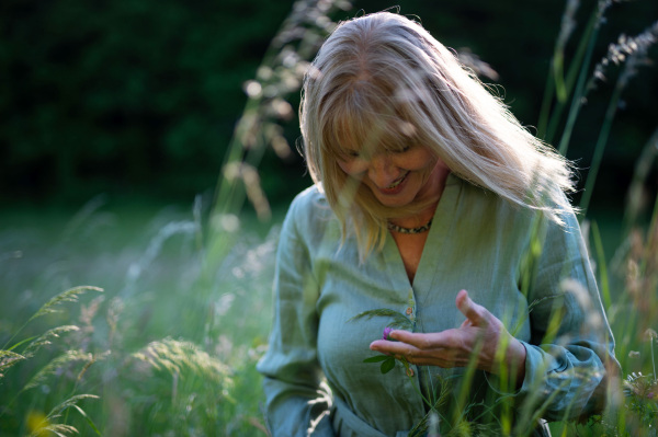 A cheerful mature woman in linen dress on walk in meadow in summer, mental health concept.