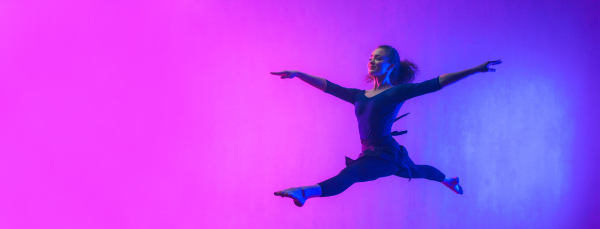 A m odern dance girl dancer jumping up dancing in neon light doing gymnastic exercises in studio, wide banner, copy space.