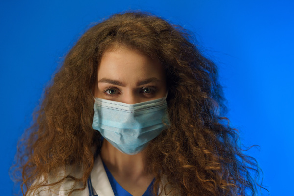 Portrait of a young female doctor with a surgical face mask looking at camera, against a blue background.