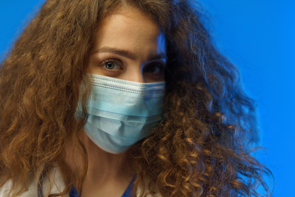 Portrait of a young female doctor with a surgical face mask looking at camera, against a blue background.