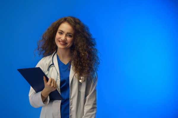 A studio shot of a young female doctor looking at camera against a blue background.