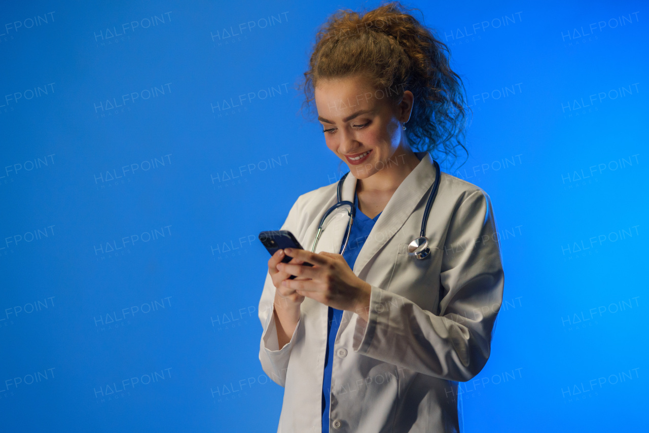 A studio shot of a young female doctor using mobile phone against a blue background.