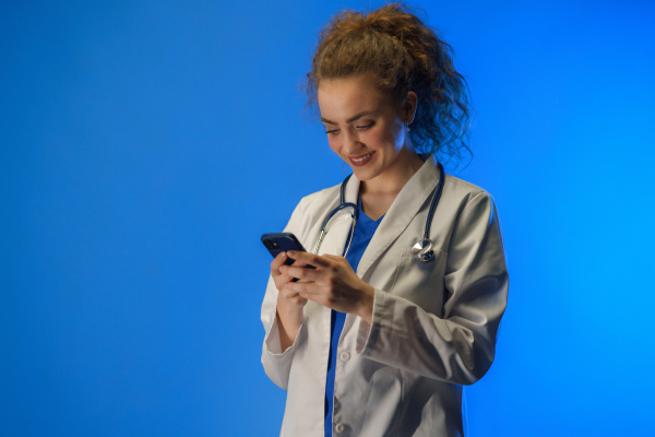 A studio shot of a young female doctor using mobile phone against a blue background.