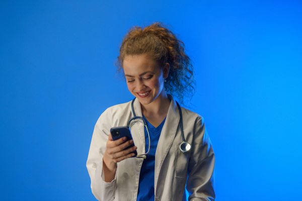 A studio shot of a young female doctor using mobile phone against a blue background.