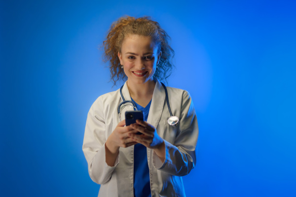 A studio shot of a young female doctor using mobile phone against a blue background.