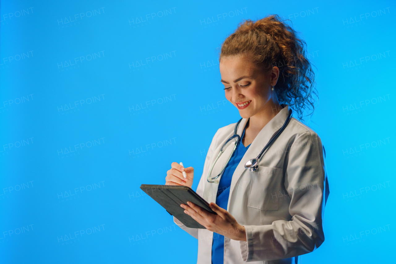 A studio shot of a young female doctor using a digital tablet against a blue background.