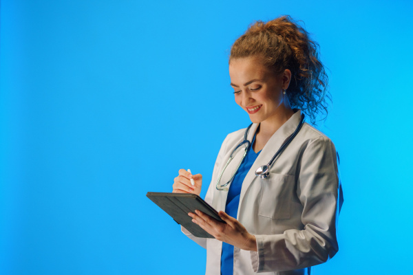 A studio shot of a young female doctor using a digital tablet against a blue background.