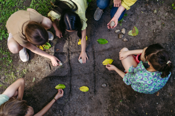 Students playing tic-tac-toe with stones and leaves, grid in soil, having fun. Outdoor sustainable education class.