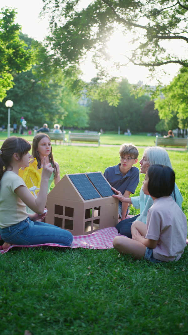 Children learning about renewable solar energy during sustainable education class outdoors, using cardboard model of house wit solar panel on roof.