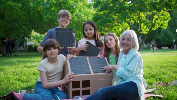 Children learning about renewable solar energy during sustainable education class outdoors, using cardboard model of house wit solar panel on roof.