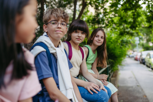 Four friends classmates with backpack sitting outdoors, waiting for bus.