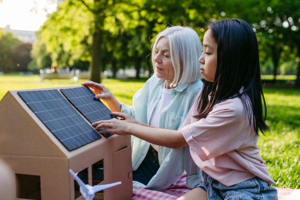 Girl learning about renewable solar energy during sustainable education class outdoors, using cardboard model of house wit solar panel on roof.