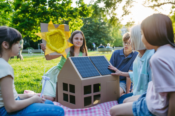 Children learning about renewable solar energy during sustainable education class outdoors, using cardboard model of house wit solar panel on roof.